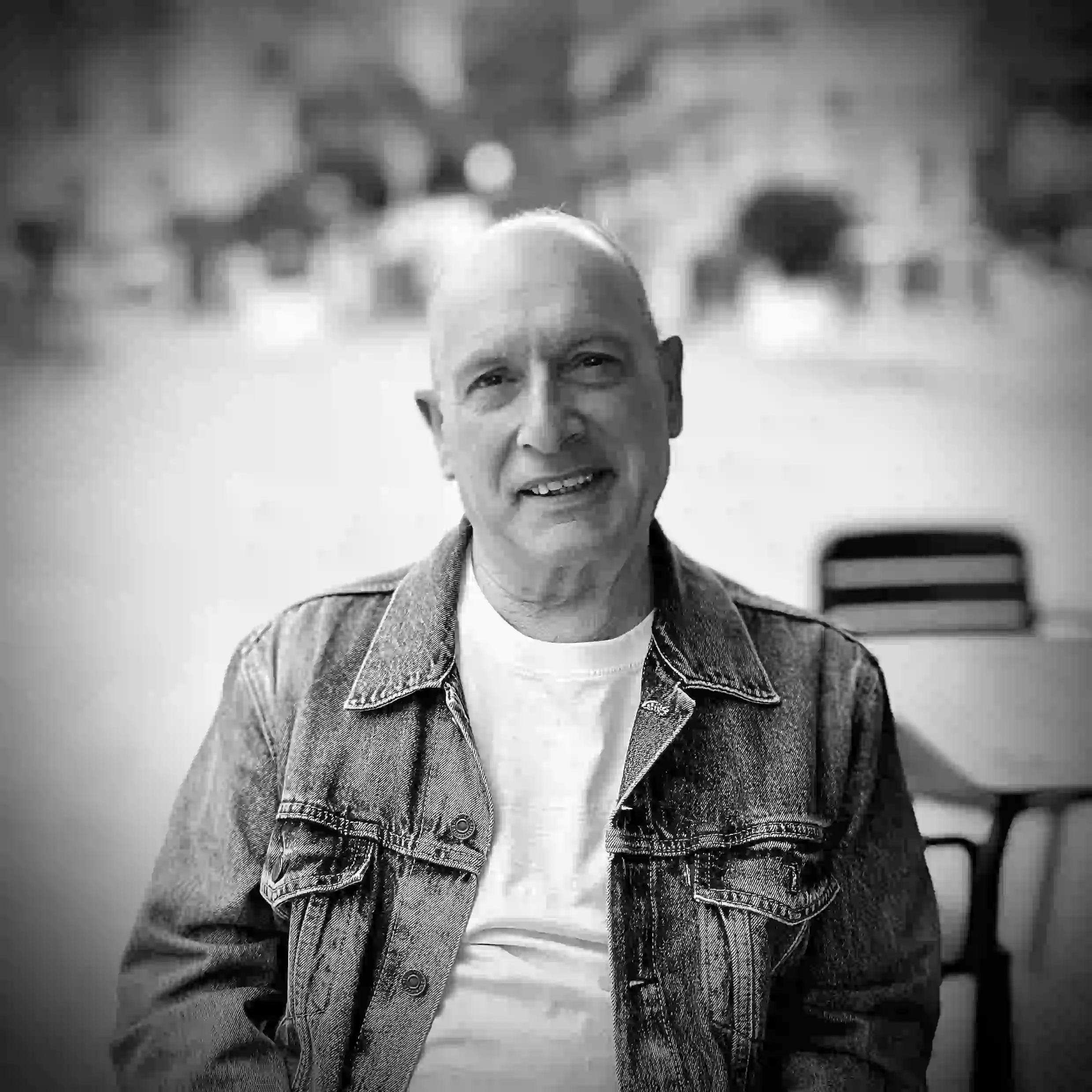 Smiling bald man, young for his age, wearing a denim jacket and white shirt sitting indoors with blurred background. Spain Valencia.